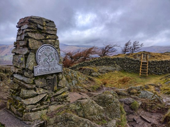 The view from Black Crag. In the foreground a pillar sits atop the rocky peak; metres away a dry stone wall is straddled by a ladder like stile. 

On the other side of the wall the trees lean to the right, reshaped by the perennial battering of the winds. The low clouds seem to hem in the landscape.

