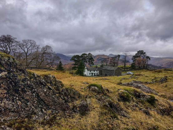 A wild looking Lakeland landscape. The rocky foreground frames the simple, white washed farmhouse in the distance. The skies are ominous, with threatening dark grey clouds as far as the eye can see.